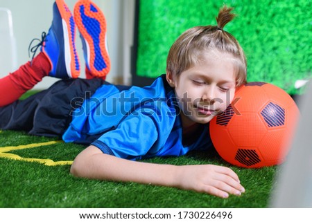 Young football player tired to play at home on green field near TV screen during the quarantine. Stay at home. Sport activities. Coronavirus situation. Sleeping boy. Kids activities.