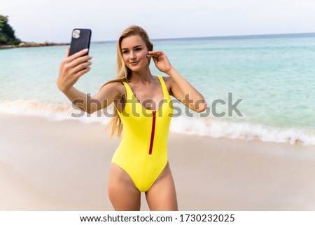 Smiling female tourist in tropics enjoys free time outdoor near ocean on beach. Looks at camera during leisure on summer day. Poses for selfie