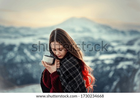 Young brunette girl drinks hot tea in the winter mountains of the Caucasus. The girl wrapped herself in a warm plaid and holds a mug of hot tea in the winter traveling in the mountains