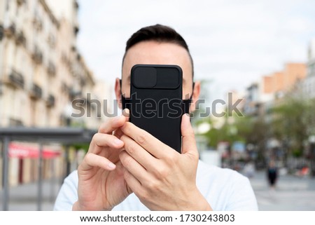 closeup of a young caucasian man on the street, wearing a black face taking a selfie or doing a video call with his smartphone, placed in front of him