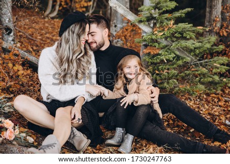 Stylish family in the autumn forest. A young guy and a girl are sitting on yellow leaves near a wooden fence with their daughter.