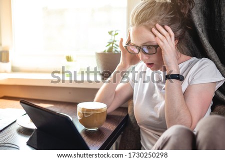 A young beautiful woman reading interesting news on the Internet using a gadget tablet sitting at a table near the window. Concept of spending the morning at home