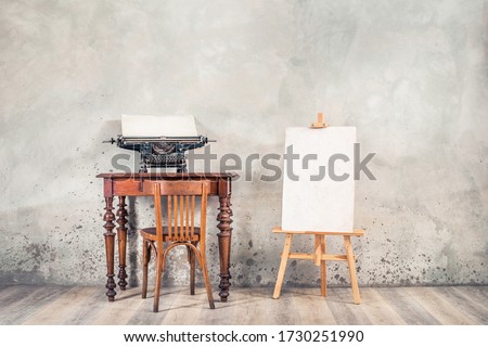 Vintage typewriter with sheet of paper on old oak desk, easel, painting canvas blank, aged chair front grunge concrete wall background. Artist's classic workplace concept. Retro style filtered photo