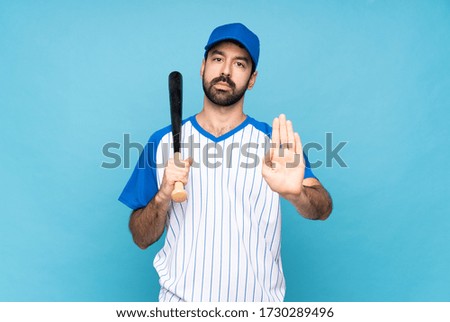 Young man playing baseball over isolated blue background making stop gesture