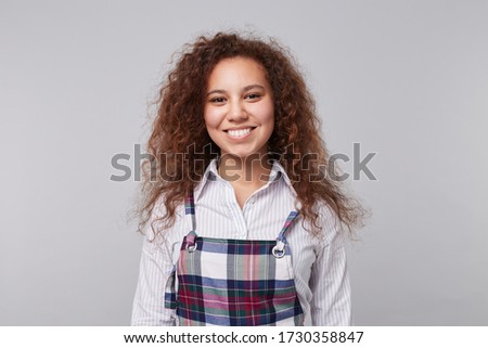 Cheerful young lovely long haired curly brunette woman showing her white perfect teeth while smiling happily at camera, standing over grey background