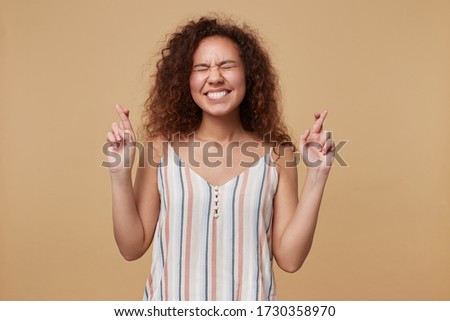 Pleasant looking young lovely brown haired curly woman keeping her hands crossed while making wish and crossing fingers for good luck, isolated over beige background
