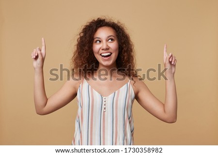 Excited young lovely brown haired curly woman in summer striped top pointing cheerfully upwards with raised forefingers and smiling broadly, isolated over beige background