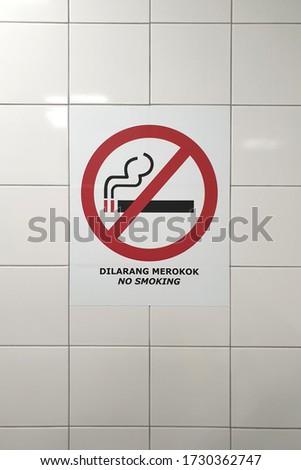 No smoking sign board in malaysian language "DILARANG MEROKOK" white and red colored board hanging on ceramic tiles wall in public toilet.
