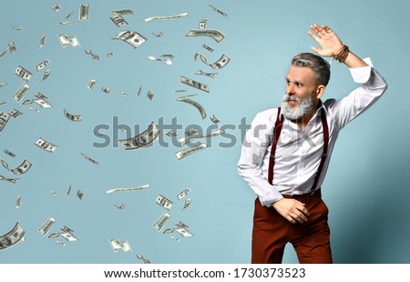 Gray-haired, aged man in white shirt, brown pants and suspenders, bracelet. He looks scared, swatting at hundred dollar bills which pursuing him, blue studio background. Close up, copy space