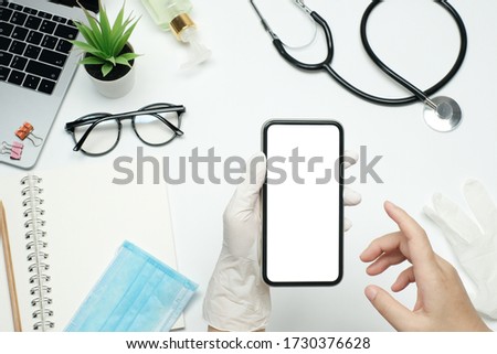 Doctor hand holding with blank screen phone on white table desk with stethoscope and sanitizer gel, medical mask and blank note pad, lab top computer and glasses