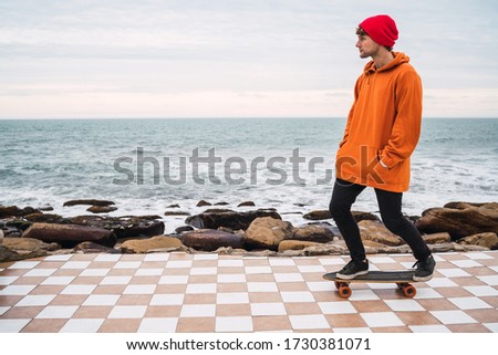Portrait of young man having fun with his skateboard and practising his tricks with sea at the background.