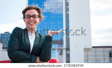 Young smiling businesswoman in glasses standing outdoors and holding copyspace on one palm. Blue sky and skyscraper glasses buildings on background. Happy office worker employee woman looks