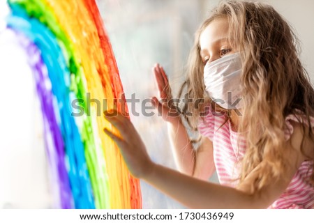 A child in a medical mask looks out the window through a painted rainbow on glass. A flash mob, a child draws a rainbow on a window pane during world quarantine due to coronavirus infection.

