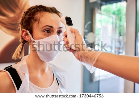 Woman checking temperature to a client using infrared tool in beauty salon - social distancing concept