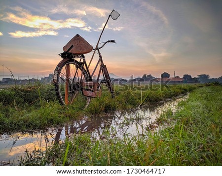 A beautiful photo of a bicycle on  dirt road that is overgrown with grass and partially inundated with water,set against the morning sky.
Location in Sukoharjo, Indonesia.
