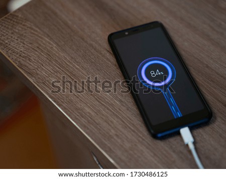 A black phone is charging at the edge of the table, a white charging cord and a blue charge indication