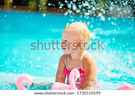 Funny blonde toddler girl swimming in the pool with pink flamingos and swim ring with water splash in the summer