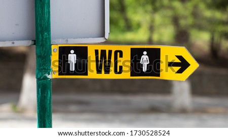 Toilet pointer, wc arrow, restroom icon, green metal sign on pillar in park.