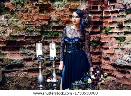 A girl in a mystical dark image in a black dress with flowers in her hair against the background of an old brick wall. Candles, black magic, Gothic beauty, mystical image. Halloween, black widow