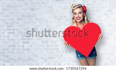 Woman holding red paper heart shape. Pin up girl. Model at retro fashion and vintage concept. White brick wall background. Copy space for some slogan or sign text. Valentine or Like symbol.