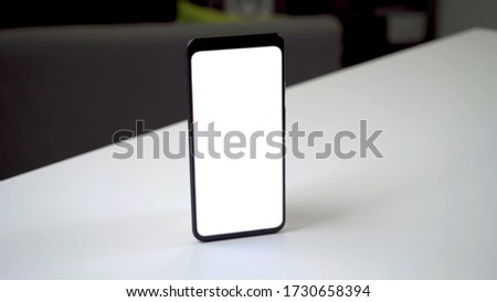 Black phone standing upright on a white table without a hand and the screen of the phone is a white template.