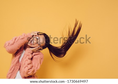 Joyful attractive brunette in glasses and headphones dressed in pink sweater white blouse shakes her head with long flowing hair while dancing, over yellow background. Positive people concept.