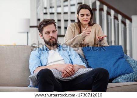 Confused man sits on the couch while his girlfriend yells and quarrels with him at home. Big family problems. Royalty-Free Stock Photo #1730712010