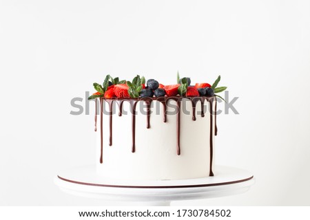 White cake with a wreath made of strawberries and blueberries and chocolate drips on the white background