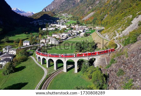 Aerial view of a Bernina Express train crossing the famous Brusio spiral viaduct of Rhaetian Railway over a green grassy meadow with fall colors on the rocky mountainside, in Graubunden, Switzerland Royalty-Free Stock Photo #1730808289