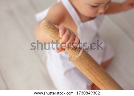 Little chef in apron and a suit of a cook, sits on a light wooden background.  Chef's hat on his head. Close up. Rolling pin in hand. 