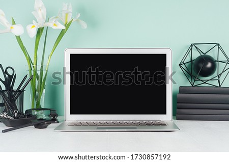Simple office interior with blank laptop monitor, black stationery, books, abstract modern sculpture, white flowers in green mint menthe interior on white wood table.
