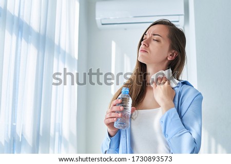 Sweating woman suffering from heat, hot weather and thirst wipes his neck with a napkin and cools down with air conditioning and cold refreshing water bottle. Refresh concept  Royalty-Free Stock Photo #1730893573