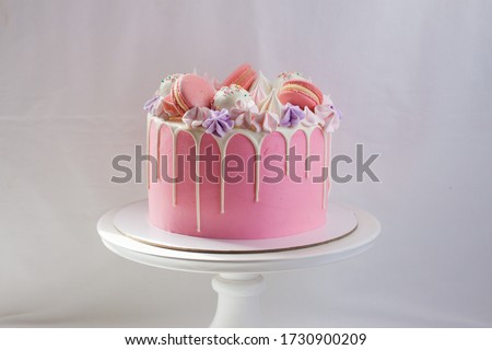 Tender pink cake decorated with melted white chocolate, macaroons, meringues, cake pops and candies on white cakestand. Plain background.