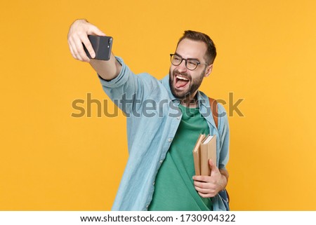 Cheerful man student in casual clothes glasses backpack hold books isolated on yellow background. Education in high school university college concept. Doing selfie shot on mobile phone showing tongue