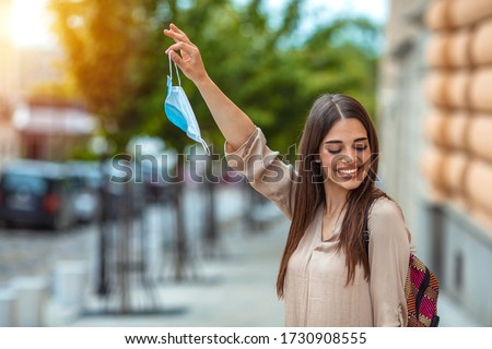 Happy young woman removing medical mask while standing in city during sunset. Woman removing protective face mask during COVID-19 pandemic. Woman throwing away her mask. Royalty-Free Stock Photo #1730908555