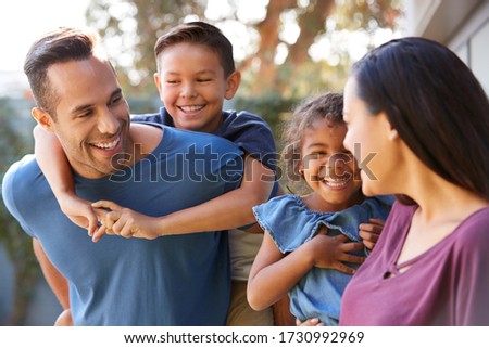 Smiling Hispanic Family With Parents Giving Children Piggyback Rides In Garden At Home