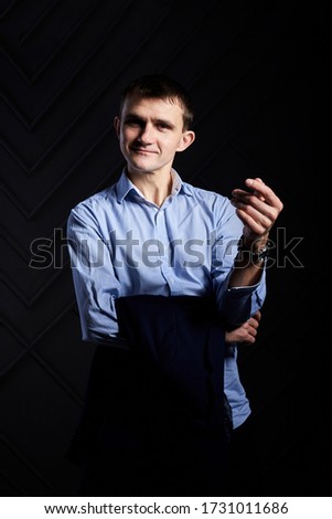 Young brunette man, wearing light blue shirt, holing his suit jacket, posing for picture in front of grey black wall in photo studio, smiling, laughing. Businessman Corporate culture. Happy Emotions.