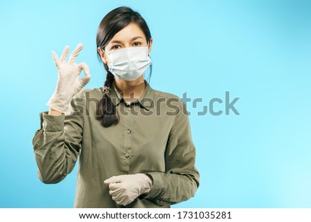 Portrait of young woman wearing a protective mask and gloves showing okay gesture on blue background. Coronavirus pandemic, dust allergy, protection against virus.
