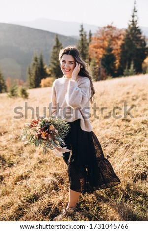 Portrait of a young beautiful girl with long hair in the autumn mountains at sunset, which holds in her hands a bouquet of flowers and greenery with a ribbon.