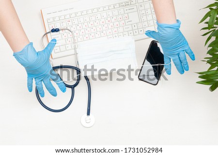 The doctor's gloved hands hold the mask. Keyboard, stethoscope, phone, mask, gloves. Top view.