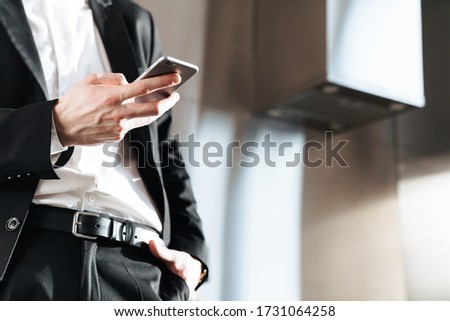 Cropped photo of caucasian businessman wearing black suit using cellphone at modern kitchen