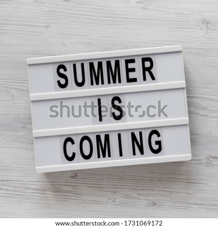 'Summer is coming' text on a lightbox on a white wooden background, top view. Flat lay, overhead, from above.