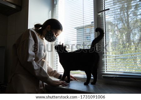 woman girl in a protective black medical mask of handwork, looks at a cat who stands on the windowsill, stands at home near the window with during quarantine covid-19, quarantine self-isolation
