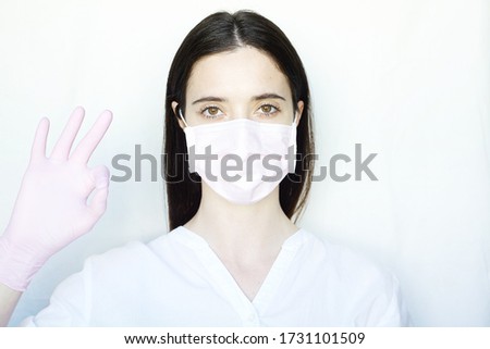 A woman in a pink protective mask and pink gloves shows a ok sign. The girl stands on a light background in white clothes. The concept of suppression of coronavirus