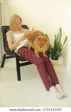  Beautiful blonde girl reads in armchair on light background. Woman without makeup at home.Portrait of happy girl with pretty pomeranian spitz in home interior.Quarantine coronavirus concept