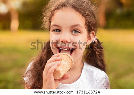 Delighted little girl with curly hair licking sweet ice cream and looking at camera while resting in park
