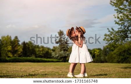Side view of sisters in similar dresses hugging and kissing each other while resting in park on sunny summer day