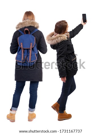 Back view of two young woman photographed on a mobile phone in winter jacket. Rear view people collection. backside view of person. Rear view. Isolated over white background.