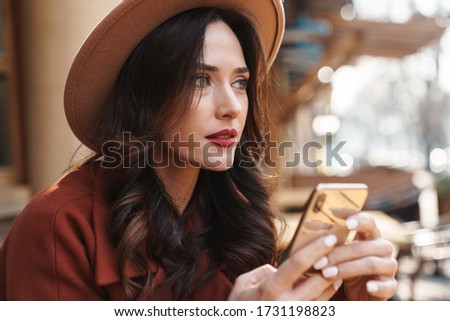Image of focused elegant adult woman in hat using mobile phone while sitting at street cafe outdoors
