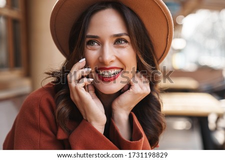 Image of joyful elegant adult woman in hat smiling and talking on cellphone while sitting at street cafe outdoors
